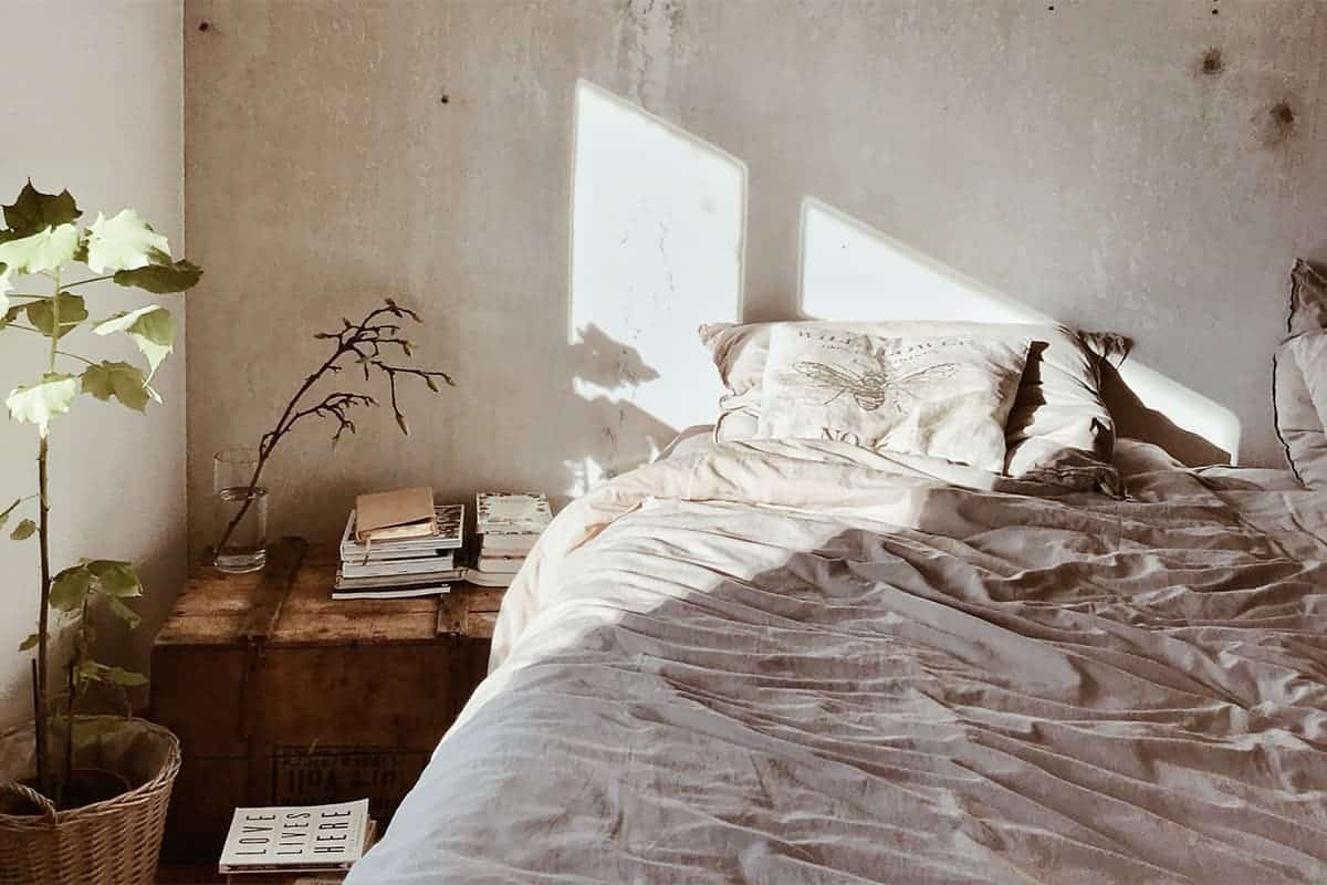 Gemütliches Bett mit grauer Bettwäsche im industrial Style neben Pflanzen und Holzkiste