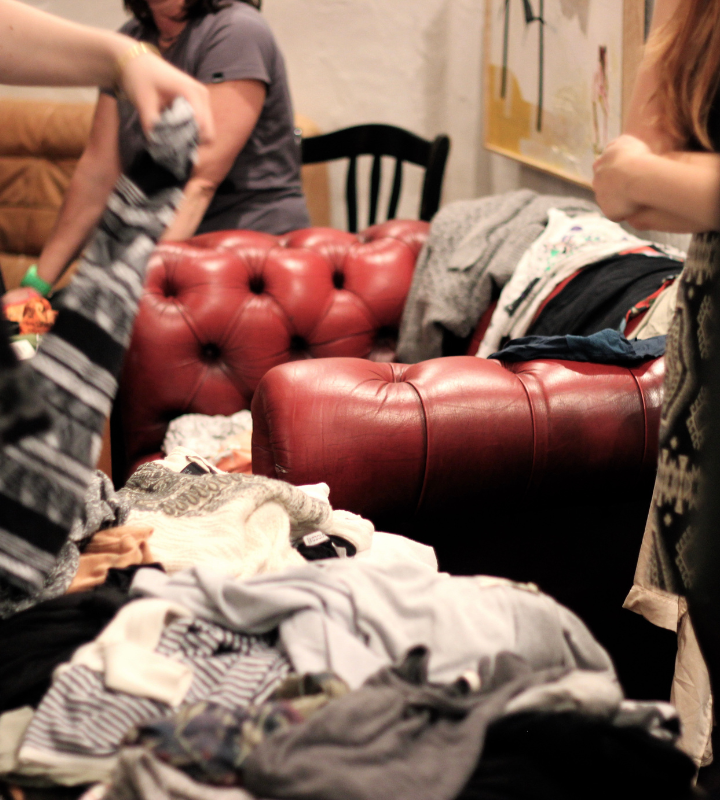 Ein Flohmarkt, ein rotes Ledersofa steht im Hintergrund, es ist bedeckt mit Kleidung. Im Vordergrund stehen Personen mit Kleidung in der Hand.