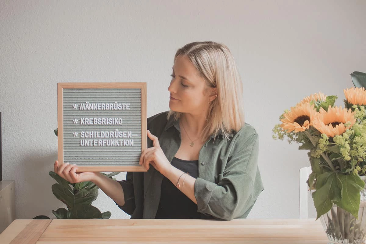 Frau an Holztisch mit Sonnenblume und Letterboard mit Aufschrift "Männerbrüste" "Krebsrisiko" "Schilddrüsenunterfunktion"