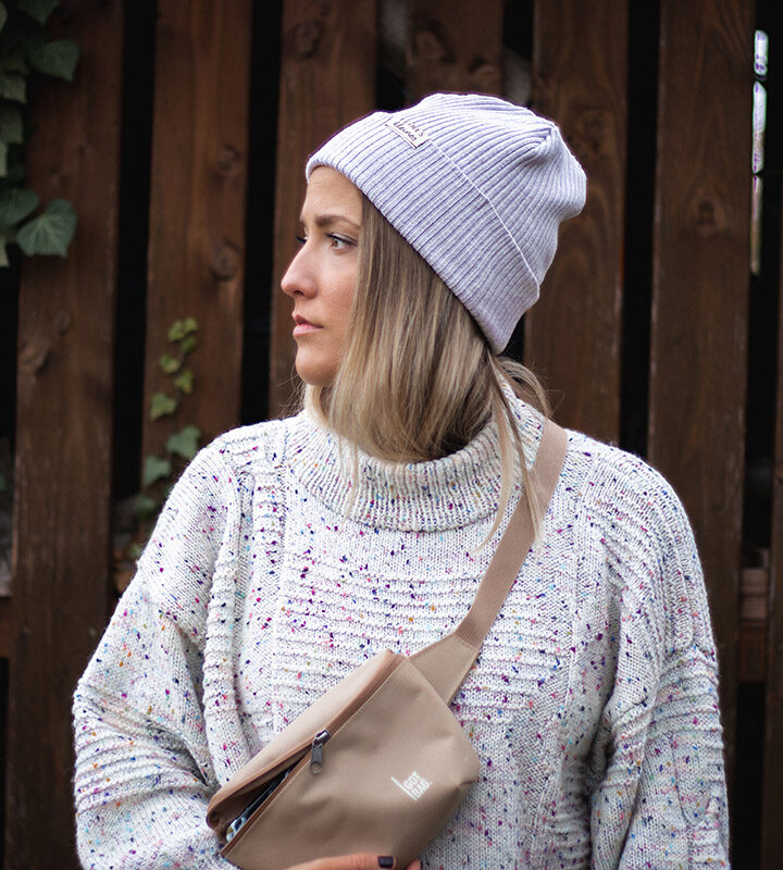 Blonde Frau mit beiger Mütze und Bauchtasche vor Holzwand