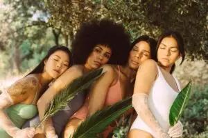 Frauen in Badeanzügen stehen vor einem Baum, halten Zweige von Pflanzen in der Hand