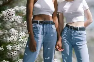 Zwei Frauen halten Hände, tragen Jeans und weiße Shirts, stehen vor weißen Blumen