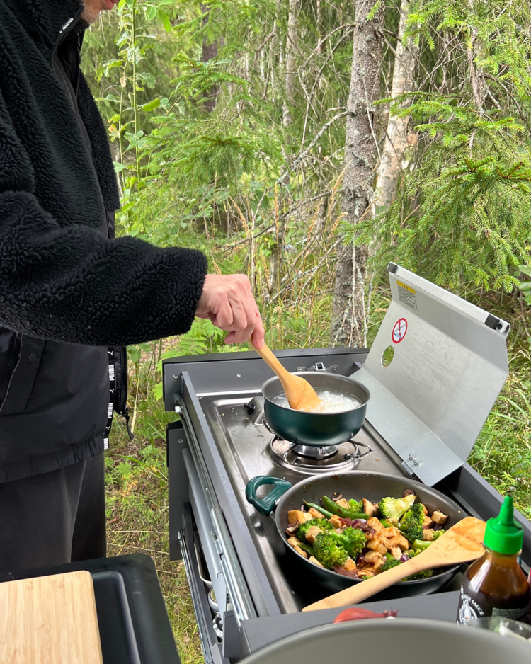 Eine Campingküche zum Ausziehen aus dem Kofferraum. Zwei Töpfe stehen darauf, ein Mann rührt in einem Topf Nudeln um. Im Hintergrund ist Wald.