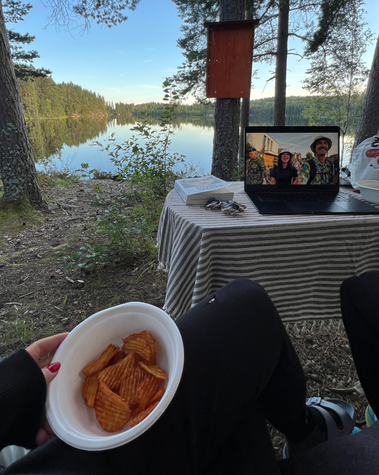 Ein POV von einer Frau, die eine Einwegpapierschale mit Chips befüllt hält, der Blick ist auf einen See gerichtet, vor der Frau steht ein Tisch mit einem Tablet darauf.