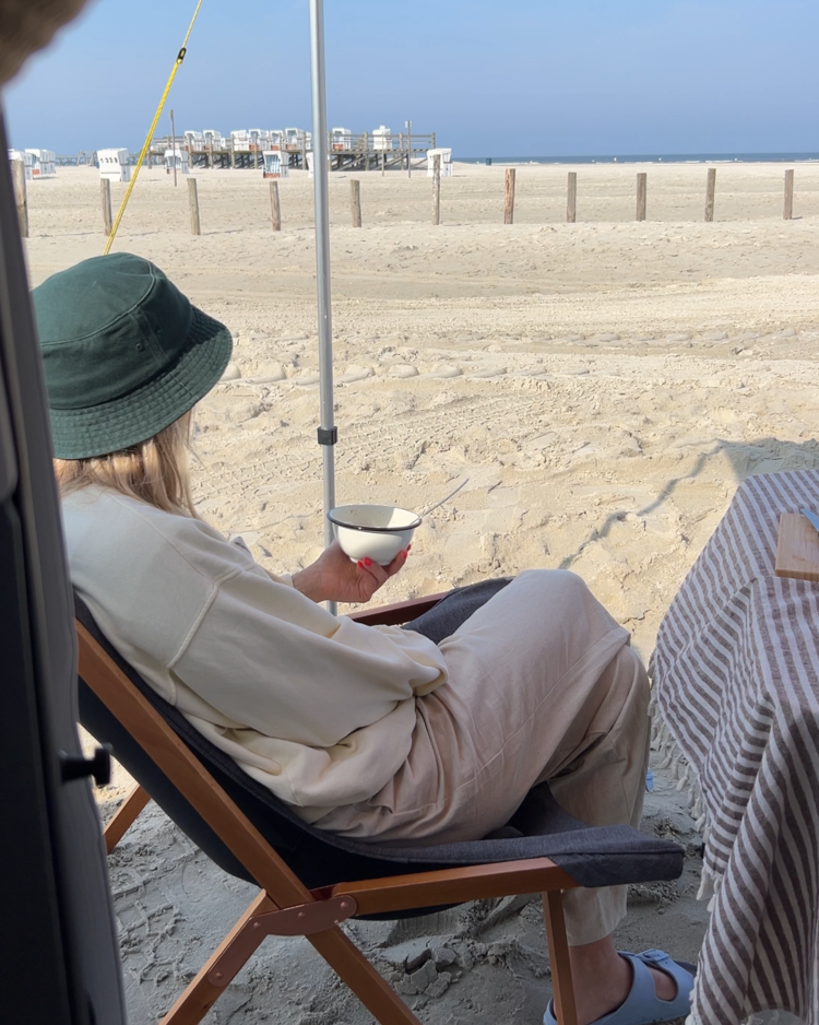 Eine Frau sitzt am Strand auf einem Campingstuhl, blickt in Richtung Meer. Sie hält eine Emaille-Schüssel in der Hand, in der ein Löffel ist.