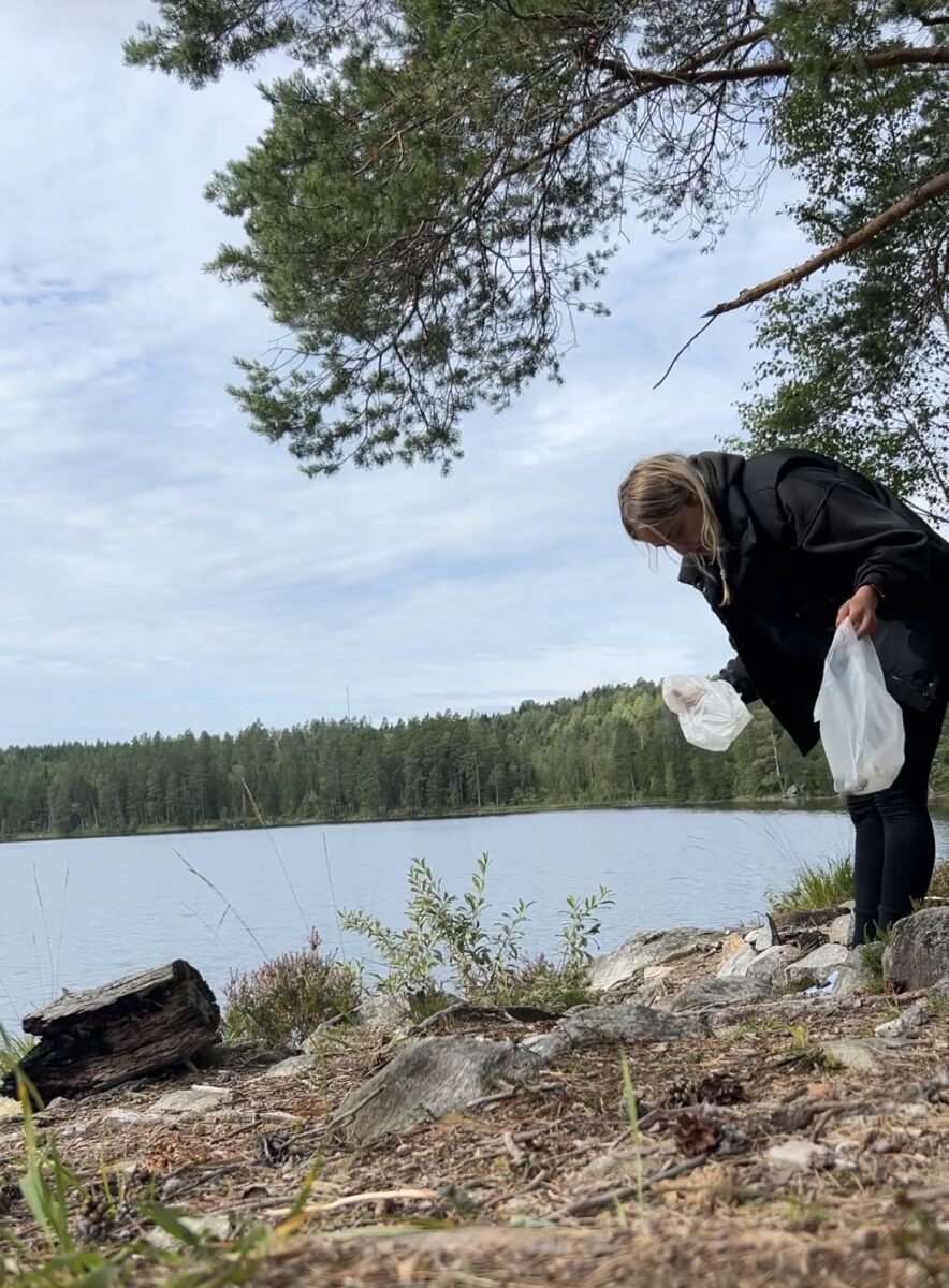 Blonde Frau mit zwei Mülltüten in der Hand, sie schaut auf den Boden und sammelt Müll auf. Im Hintergrund ist ein See und Wald.