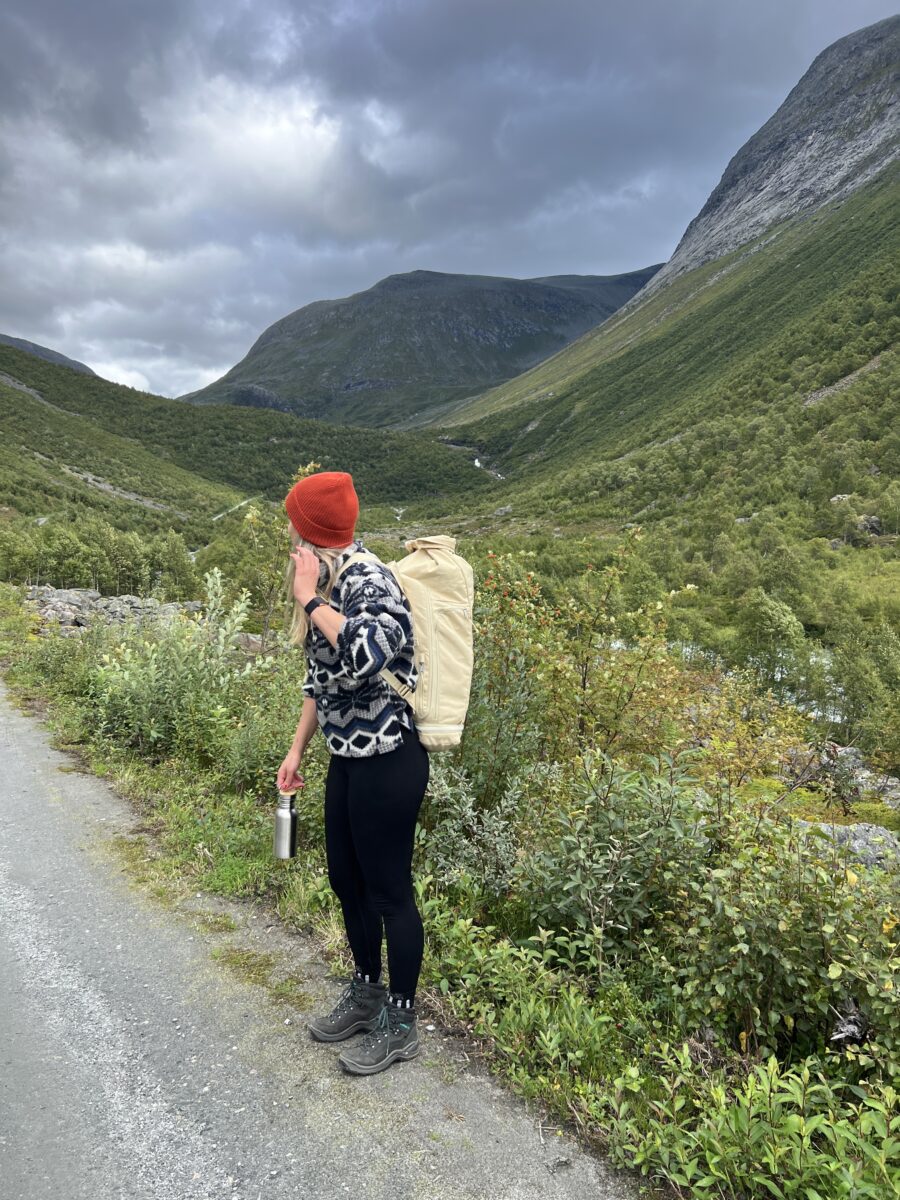 Blonde Frau trägt einen WAYKS Rucksack und eine Edelstahl Trinkflasche. Sie steht auf einem Weg. Im Hintergrund ist eine satte grüne Landschaft.