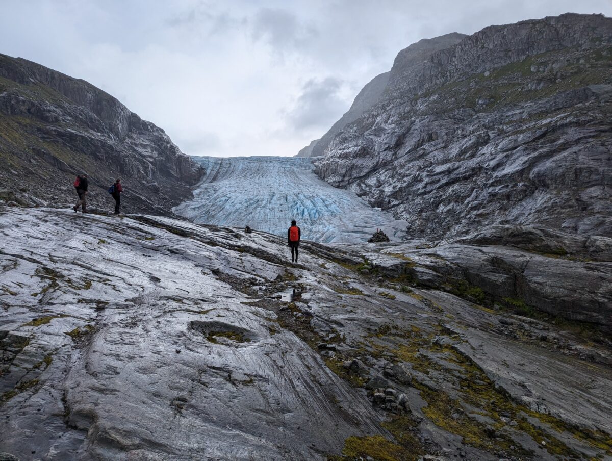Frau steht in einer mächtigen Felslandschaft, sie trägt einen Rucksack mit rotem Cover, der Rest ist grau. Vor ihr ein blauer Gletscher. Es ist der Haugabreen Gletscher in Norwegen.