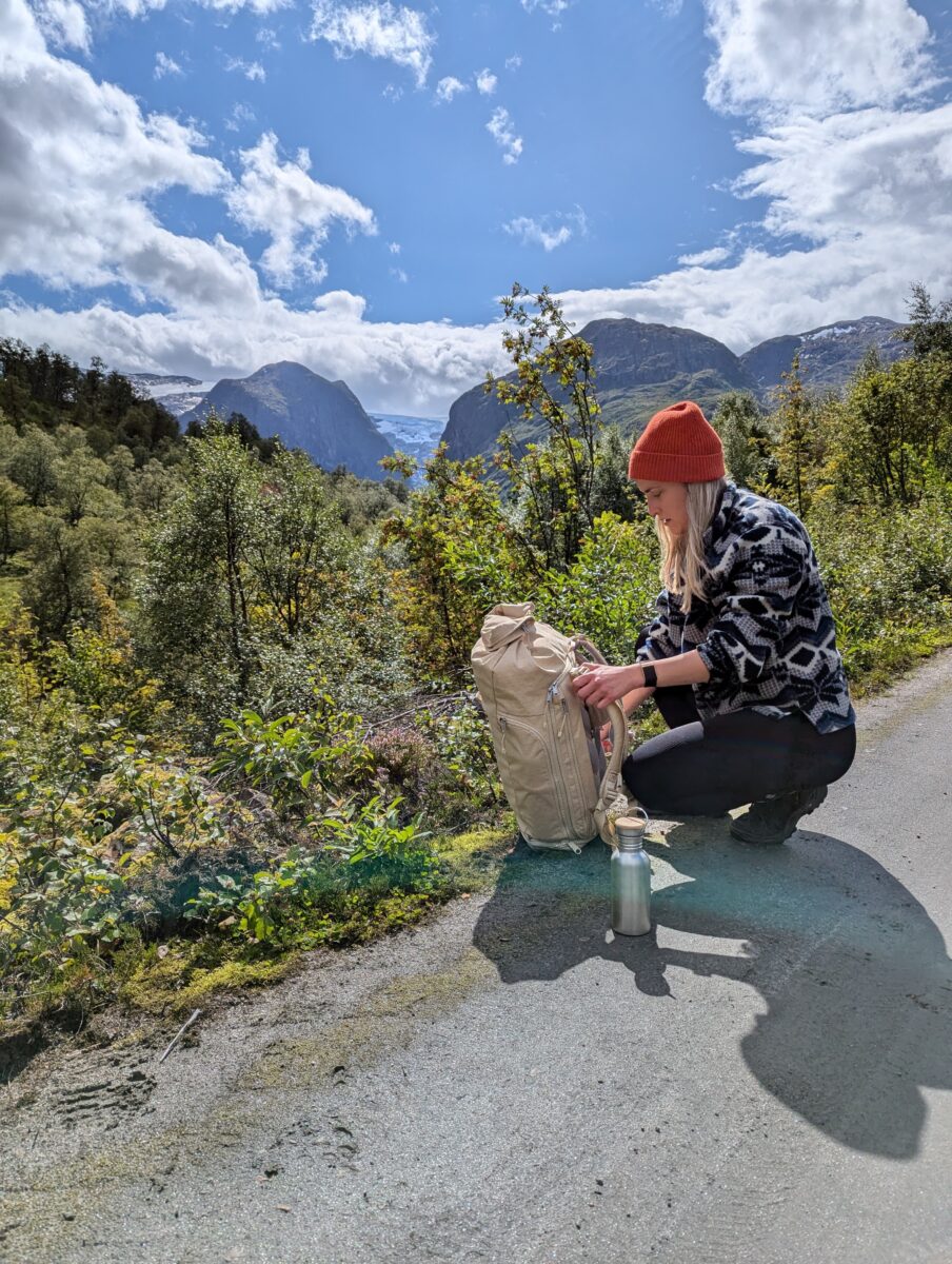 Frau kniet vor WAYKS Rucksack, neben ihr steht eine Edelstahl-Trinkflasche. Im Hintergrund satte grüne Landschaften und Berge.
